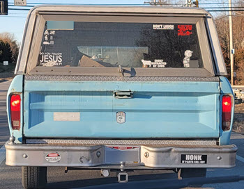Rear end of an old blue truck with covered with a cap and several bumper stickers.